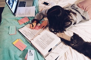 Mladá žena leží na posteli s hŕbou poznámok a papierov a pracuje na dokončení svojej diplomovej práce