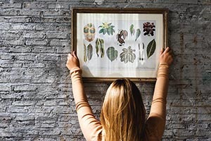 Dievča vešia na kamennú stenu zarámovaný obraz s potlačou rastlín